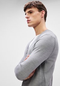 Siv šank z V-izrezom, gladka tekstura in gumb zapiranje. Prilagojen dizajn z dolgimi rokavi in rebrastimi poudarki ob robovih.