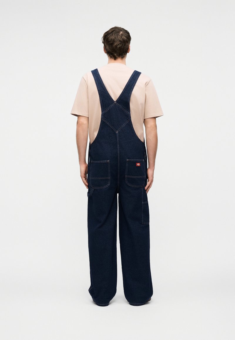 Mann, der mit dem Rücken zur Kamera steht, trägt ein hellbeiges kurzärmeliges Hemd und dunkelblaue Jeans-Latzhose mit weitem Bein vor einfarbig weißem Hintergrund.