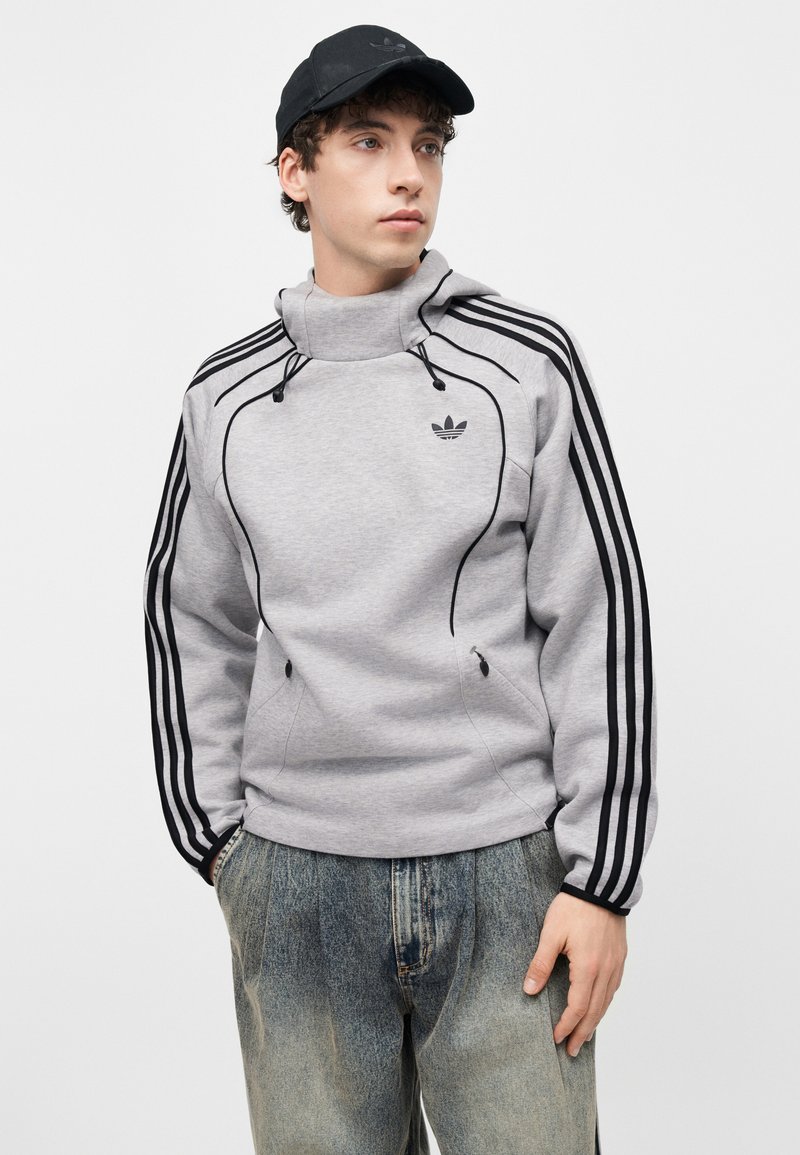 Jonge man die een grijze Adidas trui draagt met zwarte strepen en logo, zwarte pet en vervaagde blauwe spijkerbroek, kijkt opzij.