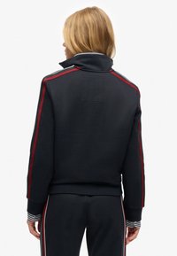 Donkerblauwe sweatshirt met hoge kraag, rood-witte strepen langs de schouders en geribbelde manchetten. Het materiaal lijkt zacht en gestructureerd.