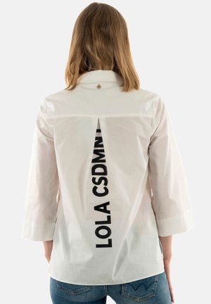 Persona con cabello hasta los hombros lleva una camisa blanca con texto vertical negro "LOLA CSDMNT" en la espalda, combinada con vaqueros azules.