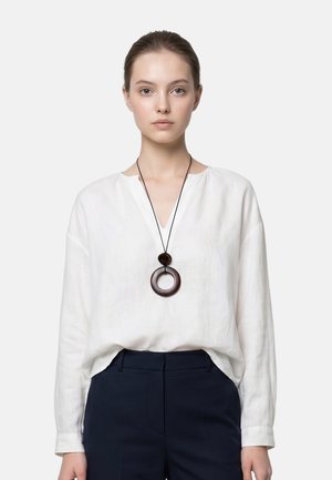 Vrouw met een lichte huid en bruin haar in een staart, gekleed in een witte blouse met lange mouwen, zwarte broek en een grote ronde hangerketting.