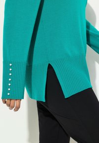 Türkisfarbener Pullover mit gerippten Bündchen und sechs Perlenknöpfen. Der Saum hat seitliche Schlitze. Kombiniert mit schwarzen Hosen, die einen strukturierten Stoff zeigen.