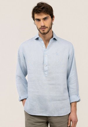 Hombre con cabello corto y castaño y barba, vistiendo una camisa azul claro de manga larga y pantalones beige, de pie con la mano izquierda en el bolsillo.