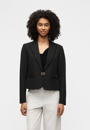 Vrouw met krullend haar, gekleed in een zwarte getailleerde blazer met gouden sluiting en lichtgekleurde broek, staand tegen een effen achtergrond.