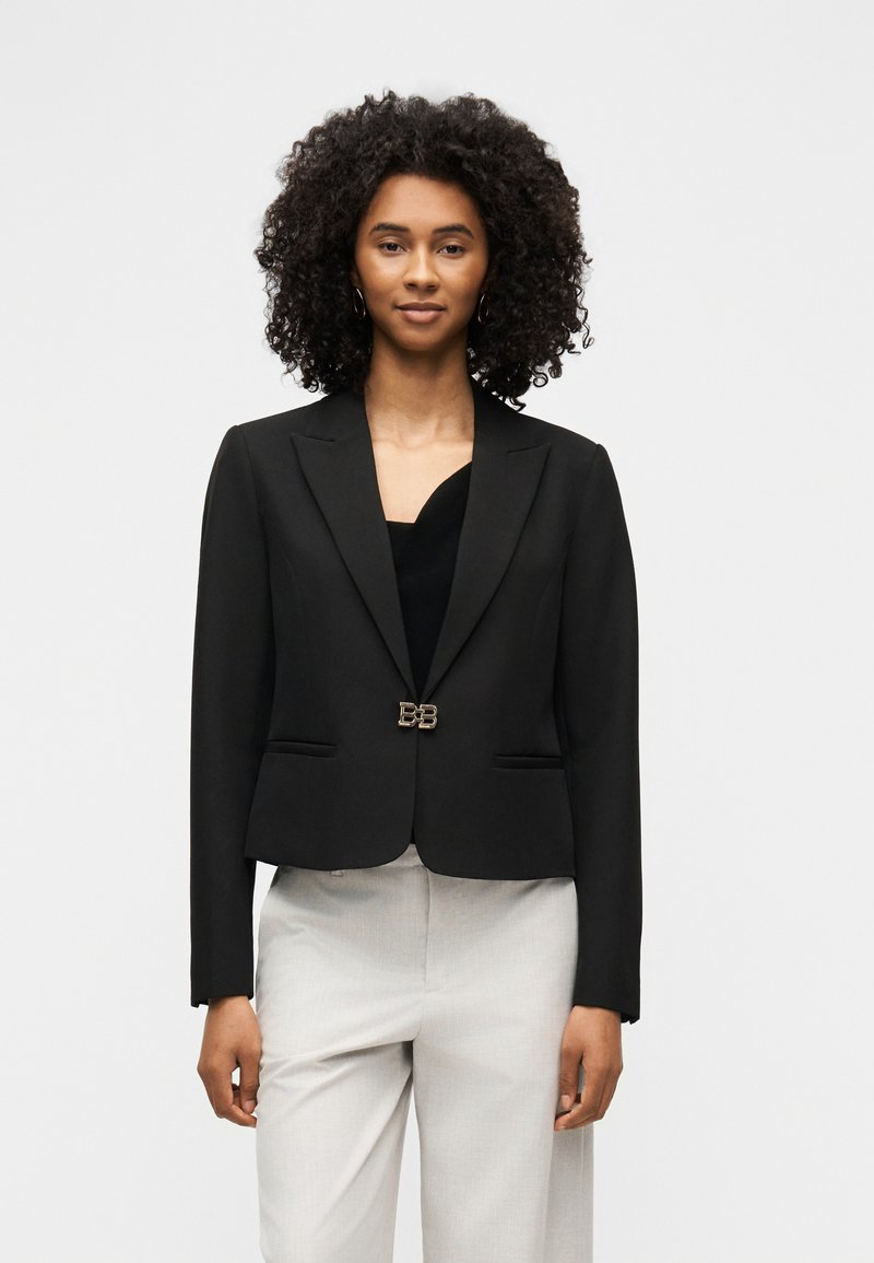 Femme aux cheveux bouclés portant un blazer noir ajusté avec un fermoir doré et un pantalon clair, debout devant un fond uni.