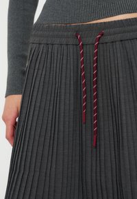 Jupe plissée gris foncé avec taille élastique et cordons bordeaux portée avec un haut gris côtelé à manches longues laissant voir le ventre.