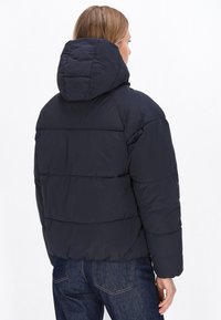 Mørkeblå pufferjakke med aftagelig hætte, der har vandrette quiltede sektioner og blød tekstur. Bæres over mørke jeans.