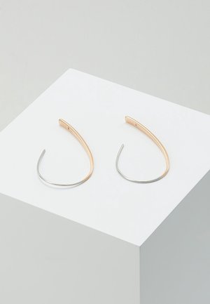 Paire de boucles d'oreilles minimalistes en forme d'anneau courbé en métal argenté et doré, présentées sur un socle cubique blanc sur fond gris clair.