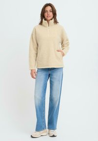 Bézs polár felső magas gallérral, félcipzáros elülső résszel és oldalzsebekkel, világoskék egyenes szárú farmerrel és bézs sportcipővel kombinálva.