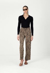 Vrouw in een zwarte lange mouwtop, met een luipaardprint broek, bruine schoenen met hakken, zonnebril en oorbellen in de vorm van een ring, staand voor een witte achtergrond.