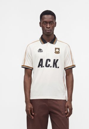 Homme portant un polo blanc avec col noir et le texte ""A.C.K."", debout devant un fond blanc uni.
