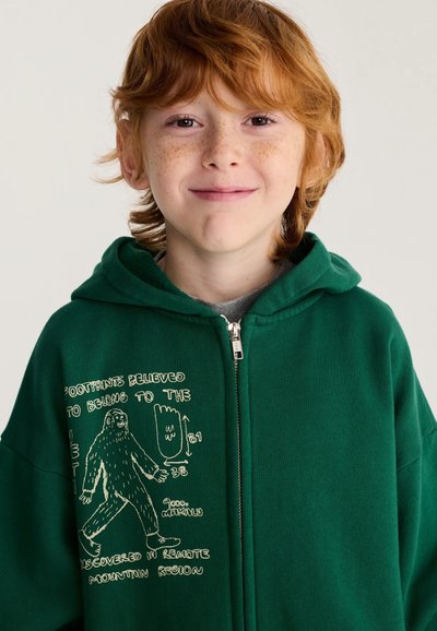 Enfant aux cheveux roux et taches de rousseur portant un sweat à capuche vert zippé avec un graphique Bigfoot blanc et du texte sur le côté gauche.
