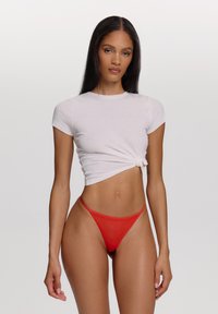 Haut court côtelé blanc à manches courtes, noué à l'avant. String rouge, couvrance minimale, avec des bretelles fines. Texture de tissu lisse.