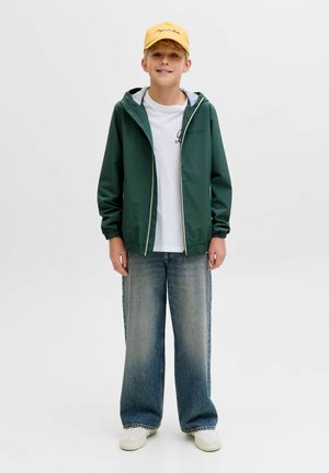 Chłopiec w zielonej kurtce na zamek, białej koszulce, szerokich niebieskich dżinsach, białych trampkach i żółtej czapce stoi na jednolitym tle, uśmiechając się.