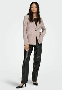 Beżowa, dopasowana marynarka na jeden guzik, zestawiona z czarnymi, błyszczącymi skórzanymi spodniami i czarnymi, szpiczastymi szpilkami. Długie, falowane czarne włosy.