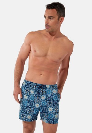 Muskularny mężczyzna w niebieskich kąpielówkach z wzorem i sznurkiem, stojący z rękami w kieszeniach, patrzący w bok.