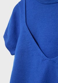 Gros plan d'un vêtement en tissu tricoté bleu présentant un détail de superposition en col en V et des manches courtes sur un fond uni.