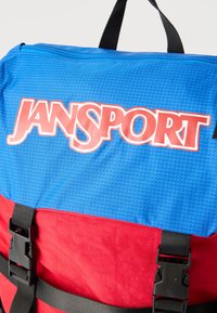 Μπλε και κόκκινο σακίδιο πλάτης Jansport με μαύρες ρυθμιζόμενες τιράντες και αγκράφες, φερμουάρ στην κορυφή, και έντονο λογότυπο στην μπλε ύφανση.