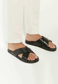 Black slide sandals with crossed straps, made of smooth leather. Flat sole, minimal design, features open toe and side foot exposure.
