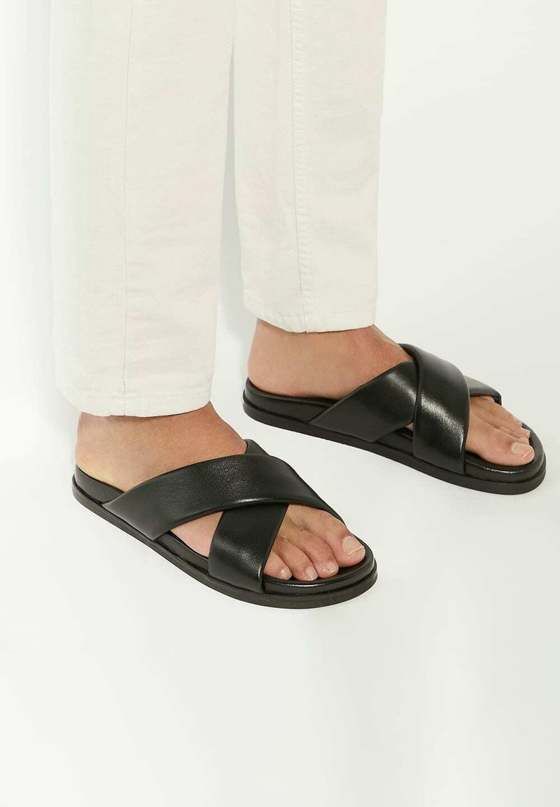 Black slide sandals with crossed straps, made of smooth leather. Flat sole, minimal design, features open toe and side foot exposure.