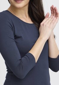 Blu închis, bluză cu mâneci lungi și decolteu rotund, textură netedă și mâneci mulate, cu detalii ușor adunate la manșete.