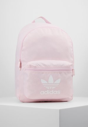 Mochila Adidas de color rosa claro con bolsillo frontal con cremallera y logo trefoil blanco, colocada sobre un pedestal blanco contra un fondo liso.