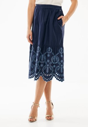 Frau trägt einen marineblauen Midirock mit bestickten floralen Ausschnitten nahe dem Saum, kombiniert mit goldenen Absatzschuhen mit Knöchelriemen und einem weißen Oberteil.