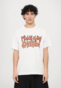 Tricou alb din bumbac, cu un text grafic îndrăzneț și unghiular, de culoare portocalie și neagră, care citește "VERSACE JEANS COUTURE". Construcție lejeră cu guler rotund.