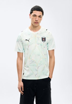 Junger Mann mit kurzem lockigem Haar, der ein weißes Puma-Fußballtrikot mit dem Logo der österreichischen Fußballnationalmannschaft und schwarze Hosen trägt, vor weißem Hintergrund posiert.