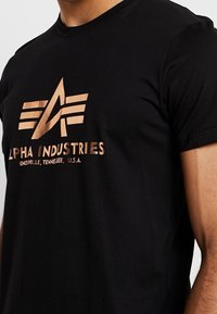 Zwarte t-shirt met een metallic koperen Alpha Industries-logo en de tekst "Knoxville, Tennessee, U.S.A." op de borst, gedragen door een persoon.