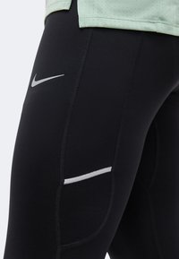 Κοντινή λήψη μαύρων αθλητικών κολαν με ανακλαστικό λογότυπο Nike και μια λευκή ανακλαστική λωρίδα στον μηρό, φορεμένα με ανοιχτό πράσινο τοπ.