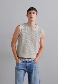 Lichtbeige gebreide mouwloze trui met een textuurpatroon, gecombineerd met lichtblauwe denimjeans en een lichtgekleurde riem.