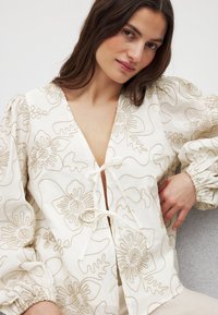 Valkoinen paita, jossa on monimutkaista beigeä brodeerausta. Syvä V-pääntie ja nyöritettävä suljenta. Puffy hihat ja teksturoitu kangas.