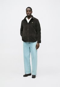 Barna kordbársony ingdzseki gombos záródással, kézelővel és zöld logóval. Világoskék, laza fazonú farmernadrág. Fekete cipő.