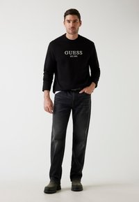 Felpa nera con logo "GUESS EST.1981", abbinata a jeans in jeans scuri e calzature nere. Design semplice, vestibilità rilassata.