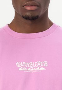 T-shirt rose à col rond en coton, avec un imprimé graphique blanc portant le mot "Quiksilver" et un slogan au centre.