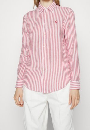 Kobieta w czerwono-białej koszuli z długim rękawem w pionowe paski, zapinanej na guziki, z małym czerwonym haftowanym logo na piersi.