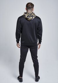 Sudadera negra con capucha estampada, que presenta diseños dorados ornamentados. Combinada con pantalones ajustados negros, que exhiben una textura suave y un ajuste atlético.