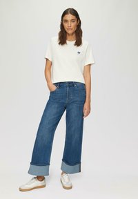Witte katoenen t-shirt met een marineblauw hartgrafiek, gecombineerd met blauwe wijde jeans met ruwe zomen en boorden, en witte sneakers met tan kleurige zolen.