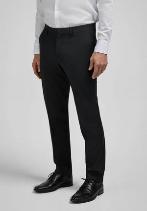 Mann trägt schwarze Stoffhose, schwarze Lederschuhe und ein weißes Hemd, steht vor einem schlichten grauen Hintergrund.