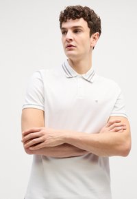 Bărbat cu păr creț purtând tricou polo alb cu guler în dungi, brațele încrucișate, privind spre stânga pe un fundal simplu și deschis la culoare.