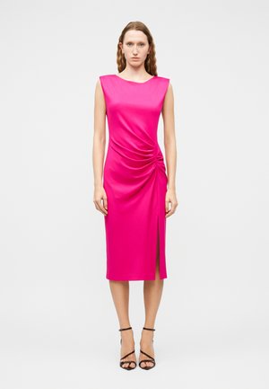 Mujer con un vestido midi sin mangas de color rosa brillante con fruncido lateral y una abertura frontal, combinado con tacones altos negros con tiras, posando sobre un fondo blanco.