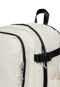 Beżowy plecak z czarnymi zamkami i paskami, z regulowanym zapięciem i teksturowanym materiałem. Boczna kieszeń z ozdobnymi detalami w postaci sznureczków.