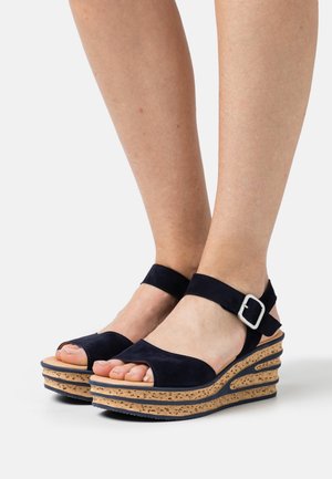 Compensées à semelles plateformes en liège, brides en daim bleu marine et boucle argentée, portées pieds nus sur un fond blanc uni.