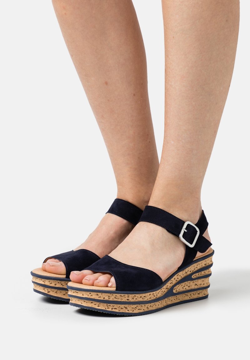 Compensées à semelles plateformes en liège, brides en daim bleu marine et boucle argentée, portées pieds nus sur un fond blanc uni.