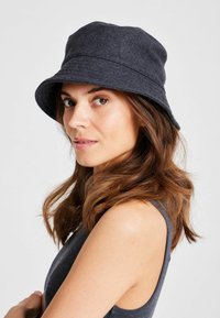 Una mujer con largo cabello castaño ondulado lleva un sombrero de cubo gris oscuro y una blusa sin mangas, mirando hacia la cámara frente a un fondo neutral.