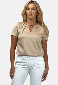 Beżowa bluzka z krótkimi rękawami, dekoltem w kształcie litery V i plisami na ramionach, zdobiona metalowym zapięciem, zestawiona z białymi spodniami.