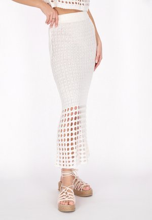 Vrouw draagt een witte gebreide midi-rok met opengewerkt onderste deel, gecombineerd met beige platformsandalen met enkelbandjes.