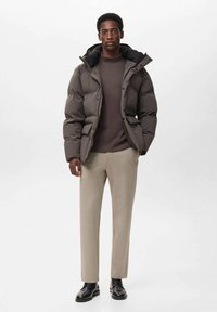 Dunkelgraue Steppjacke mit Kapuze, beige Pullover mit Rundhalsausschnitt, beiges Paar Hosen und schwarze Schuhe. Die Jacke hat aufgesetzte Taschen und eine strukturierte Oberfläche.